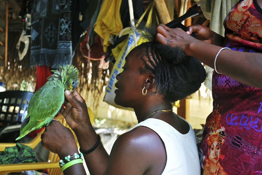 Een Surinaamse marronvrouw in Apiapaati speelt met een papegaai terwijl ze haar haren laat vlechten.