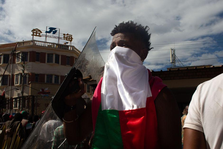 Een jongeman heeft een politieschild buitgemaakt en draagt het rond als trofee. Zijn gezicht verbergt hij met een vlag van Madagascar.