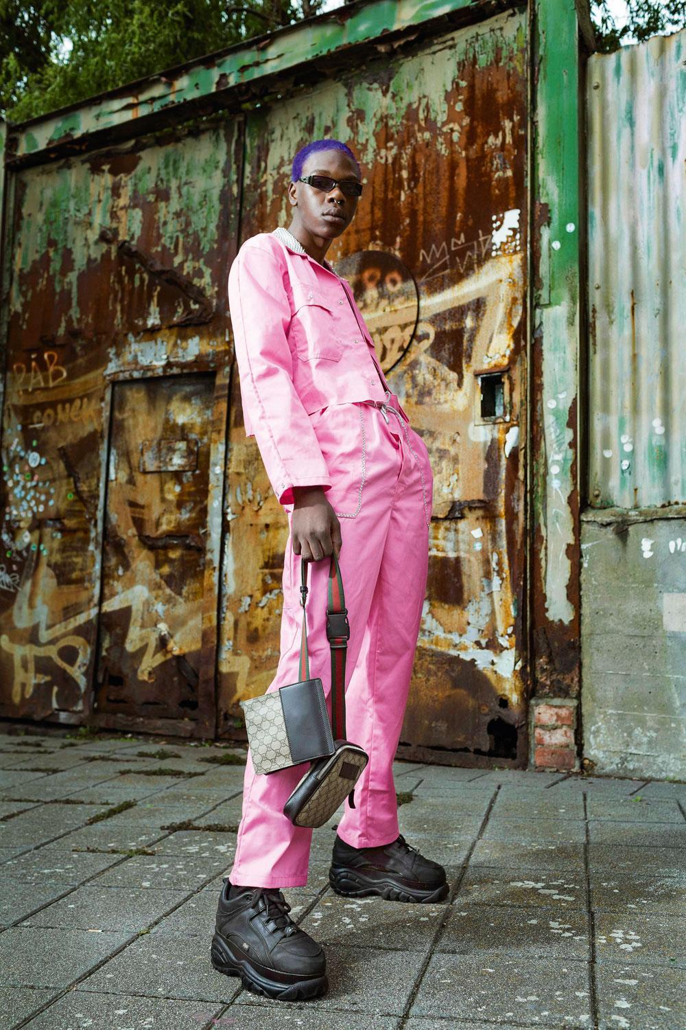 Dylan Kiala : Roze bonne suit (traditioneel werkmanspak) dat Dylan verknipt heeft, gekocht bij In 't Boerke in Antwerpen. Tweedehands Buffalo's. Gucci- tasje uit de Gucci-boetiek in Brussel