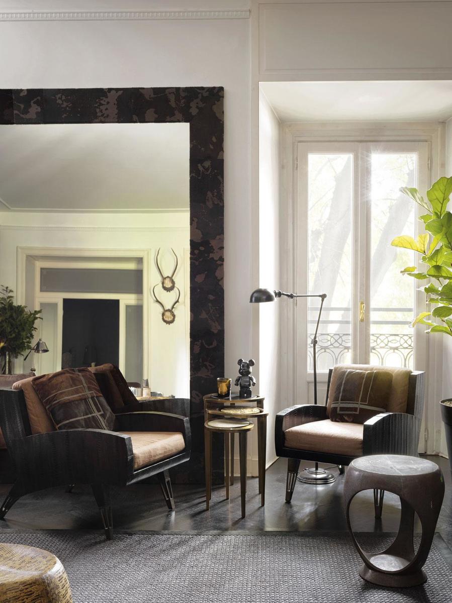 In de hoek van de zitkamer wordt een spiegel met een lijst in gezandstraalde koeienhuid geflankeerd door twee 'Saint-Germain' fauteuils en een set 'Peacock' bijzettafels van R&Y Augousti. De staande lamp en het vloerkleed zijn beide van Flamant.
