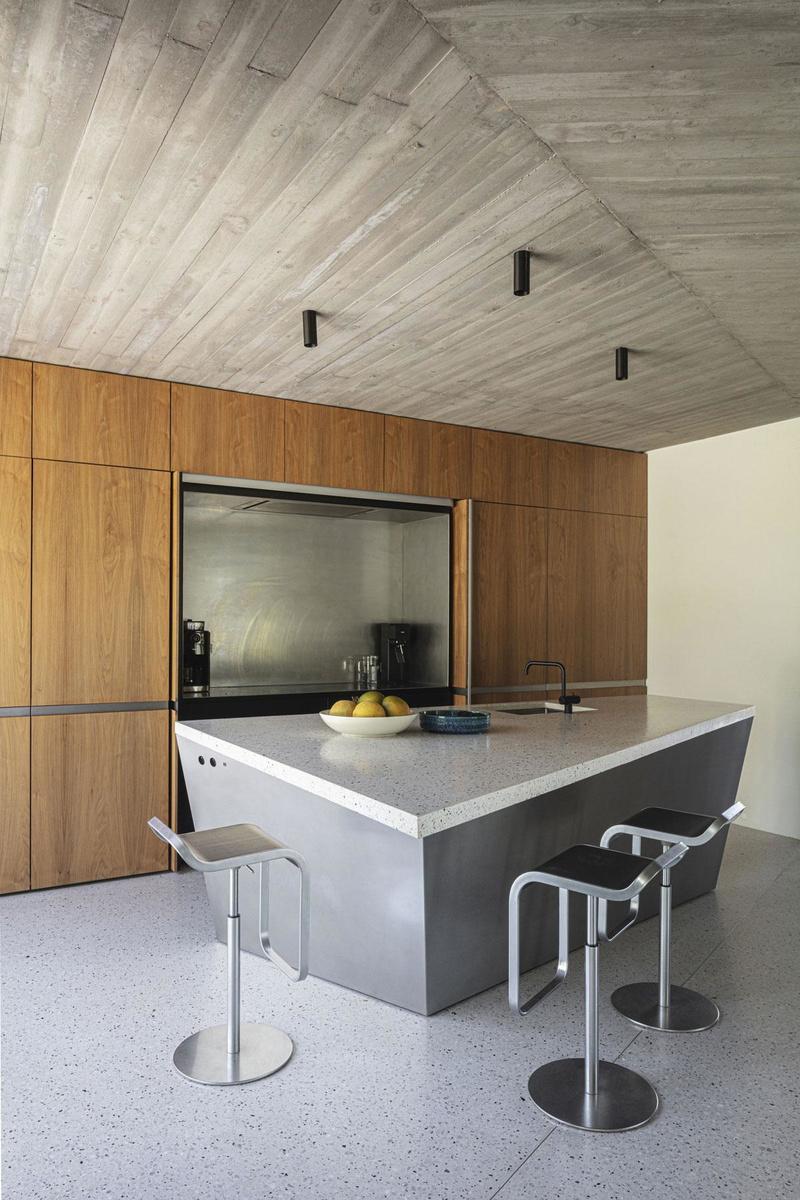 Granito, plankenbekist beton, notelaar en kathedraalglas: het materiaalpalet is beneden heel beperkt gehouden. Het asymmetrische keukenblok volgt de hoek van de ingeschoven balk, net als het plankenmotief in het plafond.