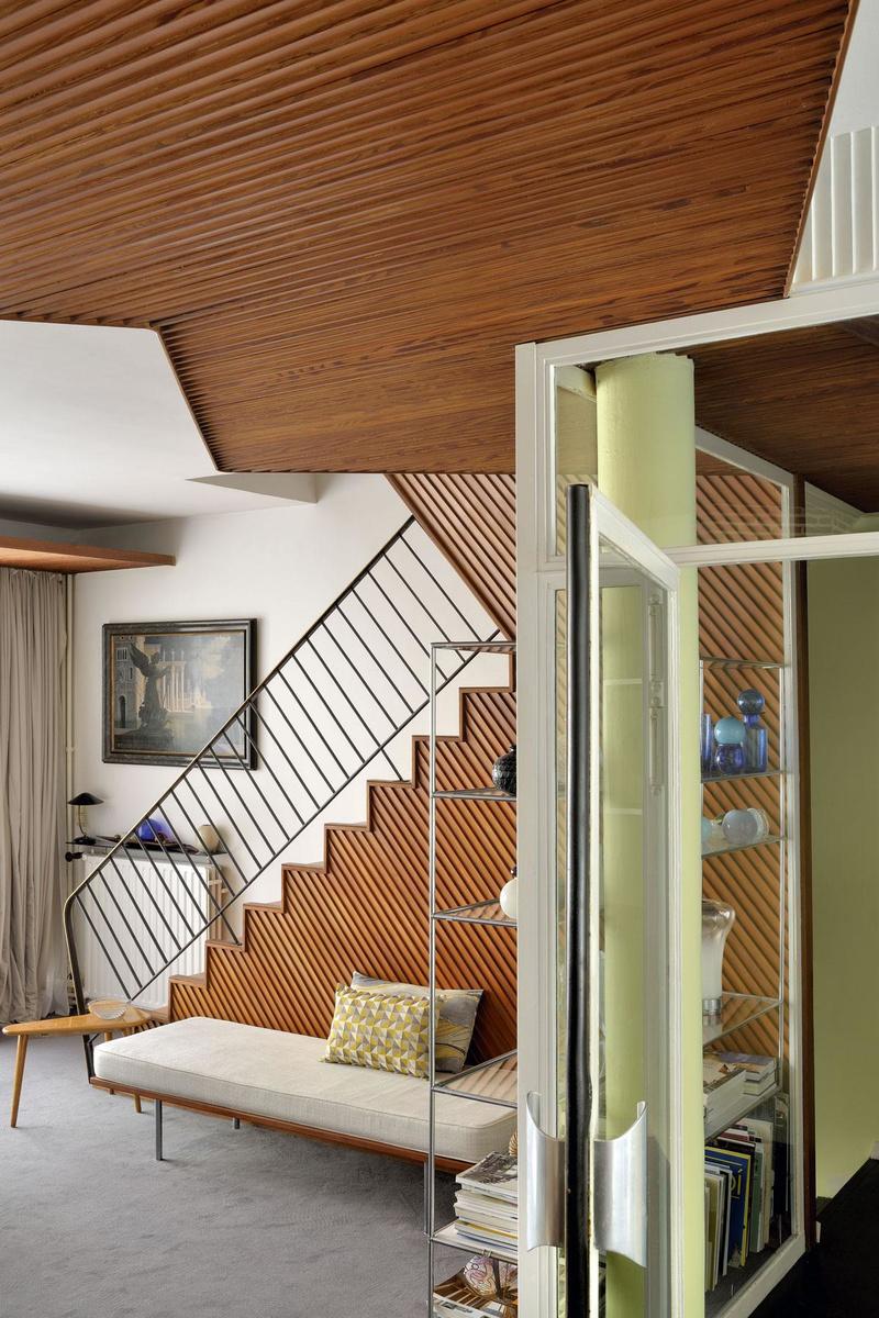 De schuine lijnen van het verlaagde houten plafond sluiten aan bij die van de lambrisering en de trapleuning, wat in de woonkamer een gevoel van dynamiek creëert.