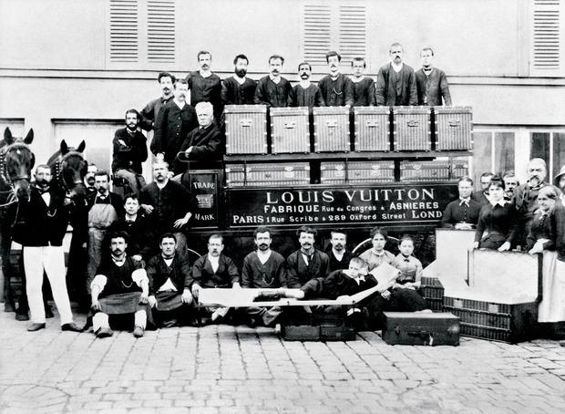 Louis, Georges et le jeune Gaston Vuitton couché sur une malle-lit, dans la cour des ateliers d'Asnières, avec leurs équipes. 