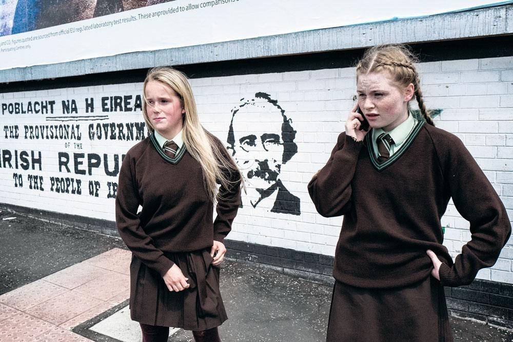 Après l'école, Tigernach (à dr.), 14 ans, et Dawn, 13 ans, attendent que le feu passe au vert à Belfast-Ouest, principalement catholique.  A l'arrière-plan, une peinture murale commémorant la proclamation de la république indépendante d'Irlande en 1916.