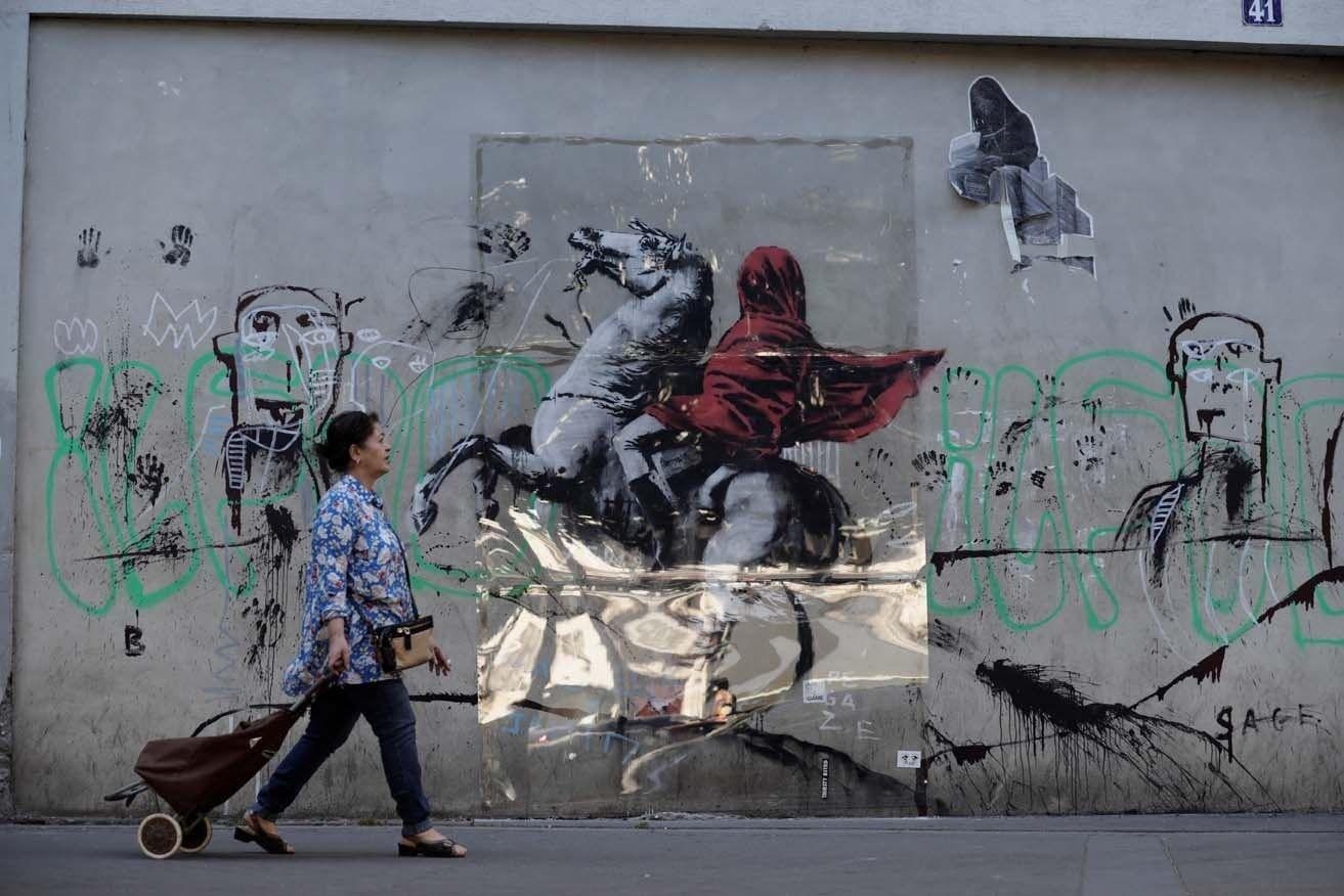 A woman walks past a recent artwork by anonymous street artist Banksy in Paris, on June 28, 2018. Following mutiple detorations of Banksy's work this week, art lovers allegedly protected with plexiglass his mural of Napoleon Bonaparte wearing a headscarf inspired by the original painting by Jacques-Louis David, in the 19th arrondissement of Paris. / AFP PHOTO / Thomas SAMSON