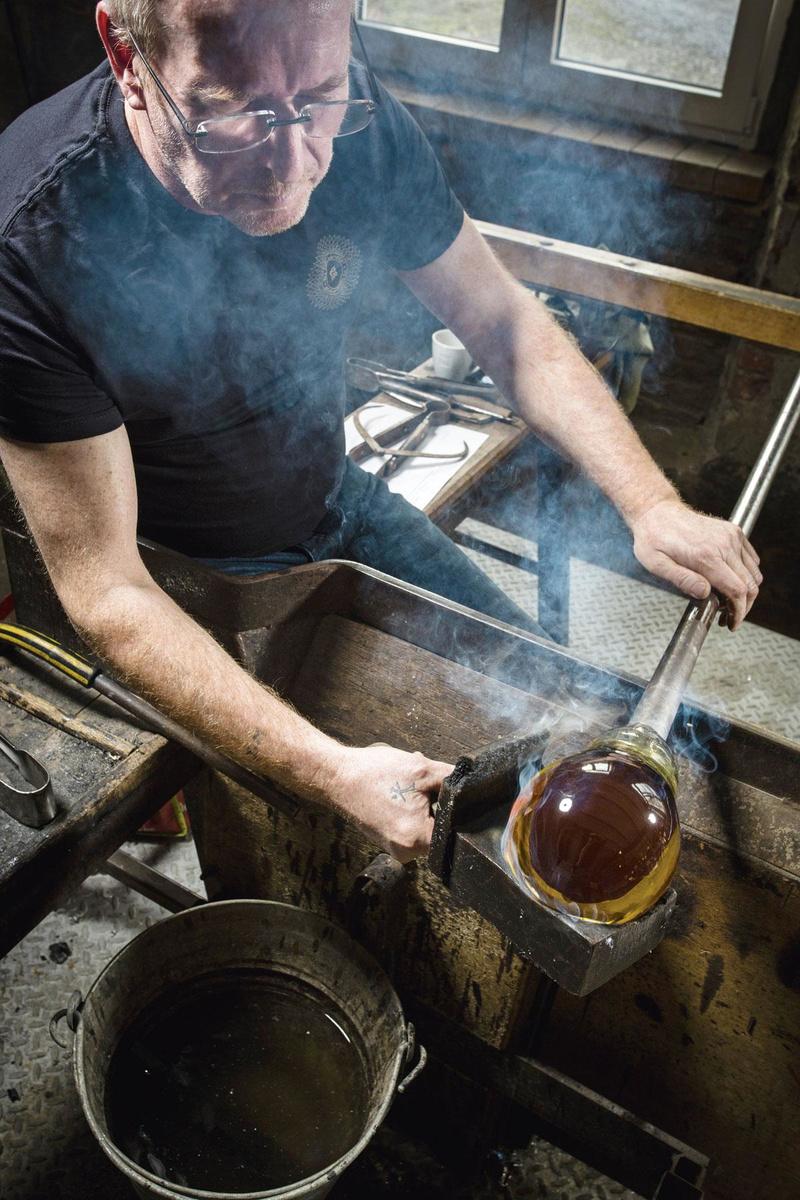 VORMGEVEN Terwijl de glasmaker blaast, laat hij  de massa vloeibaar glas onafgebroken draaien. Door die rotatie en met behulp van de klots - een uitgehold blok in perenhout - kan hij de bol kristal voortdurend opnieuw centreren en mooi homogeen en in de juiste as houden. Dat is precisiewerk, want het is zaak de bol in een as te houden die symmetrisch loopt met het gekleurde knikkertje.