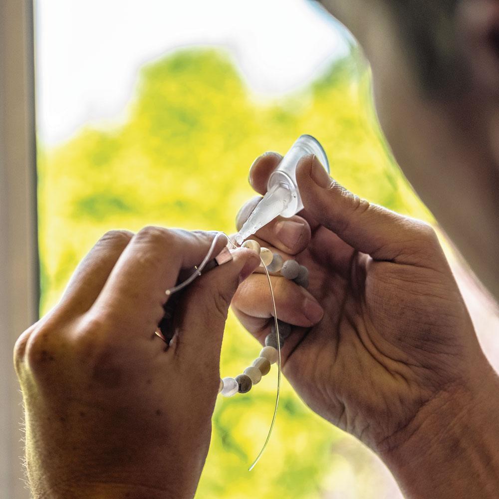 5 Lijmen - De elastiek wordt drie keer geknoopt en vervolgens zo kort mogelijk achter de knoop afgeknipt. Op het bolletje van de knoop brengt de cliënt een minimale hoeveelheid parellijm aan. De gelijmde knoop wordt daarna verborgen in een kleine holte in de armband. 