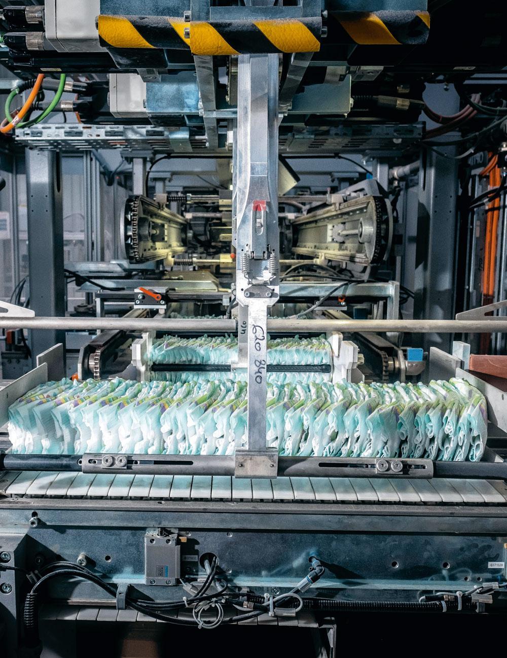 4 Découpe, pliage et collage - Le lange doit ensuite être assemblé autour de cette âme absorbante, dans une ligne de production entièrement automatisée où les rabats et les différentes couches constituant le lange sont découpés selon les formes et tailles souhaitées, puis pliés et assemblés par collage. L'ensemble du processus est en permanence sous vidéosurveillance. Les différents éléments et langes sont photographiés et comparés à un gabarit. Au moindre défaut, l'élément ou le lange est automatiquement éliminé de la ligne de production, d'où sortent environ 650 couches par minute.