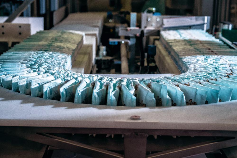 5 Double emballage - Avant l'emballage, les langes passent dans une machine qui les comprime à une pression de 2,5 à 5 tonnes, pour en réduire le volume. Tous les langes sont conditionnés dans un double emballage - d'abord dans un sac, puis dans un carton. Des robots palettisent ces cartons selon une configuration précise. Les palettes sont ensuite transportées vers la banderoleuse.