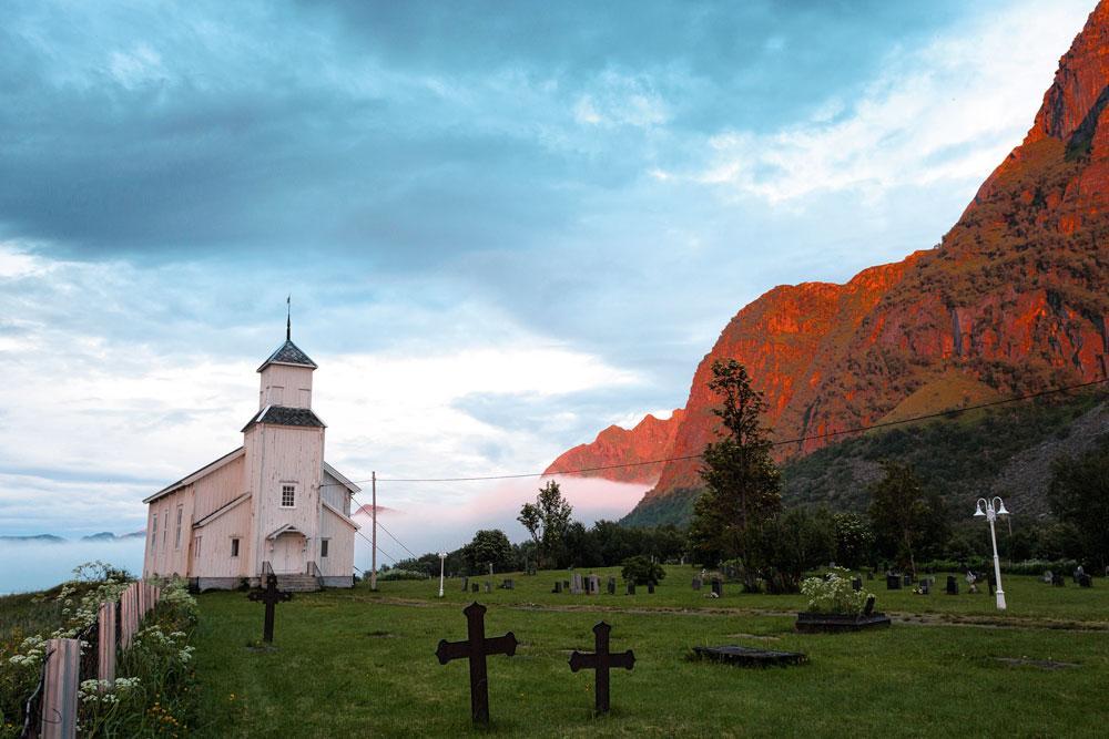 Le soleil de minuit colore les montagnes et une petite église blanche.