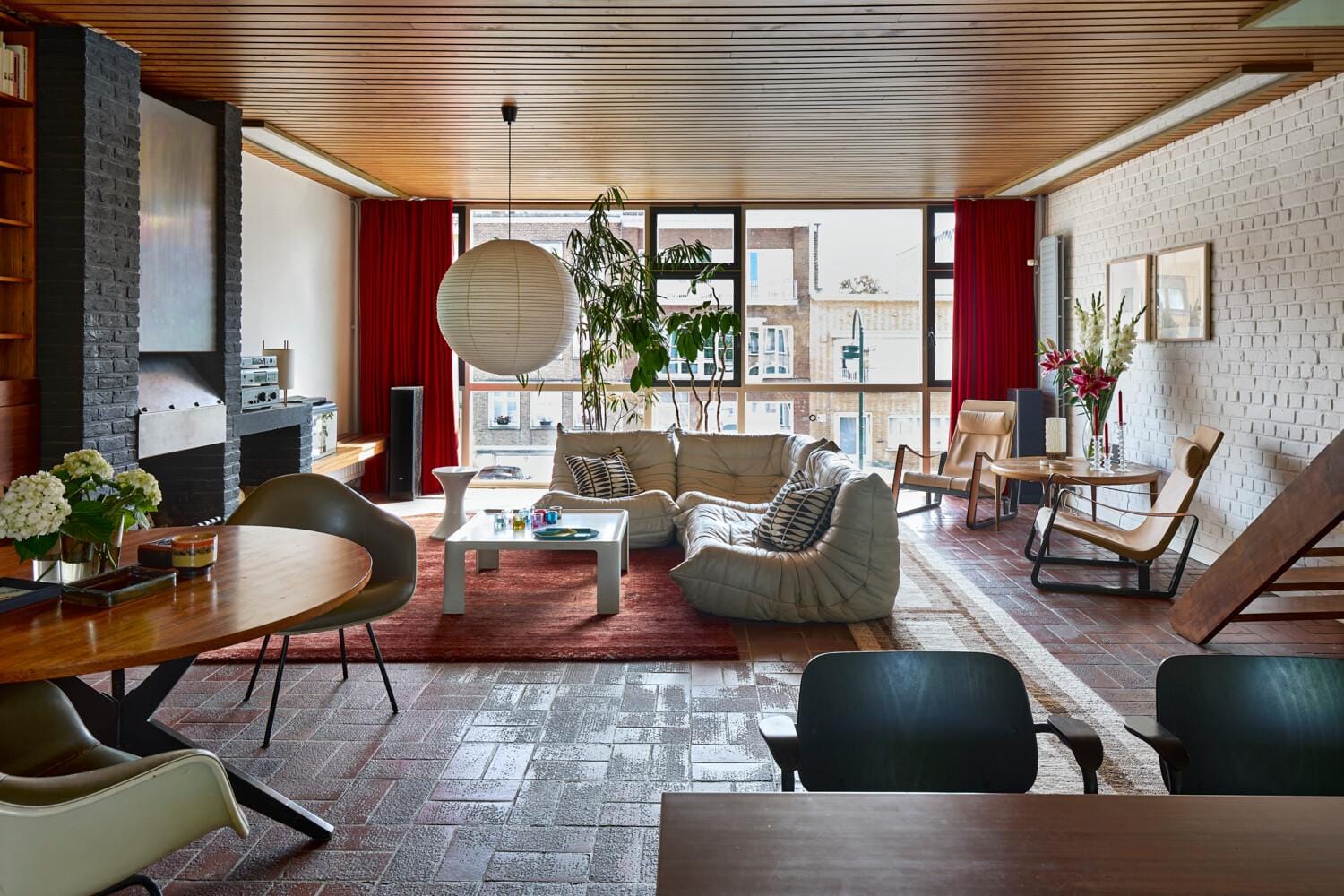 De ramen van de royale leefruimte omvatten de volledige gevelbreedte. De originele terracotta vloer, houten lambrisering en open haard met geïntegreerde zitbank zijn typisch jaren zestig.