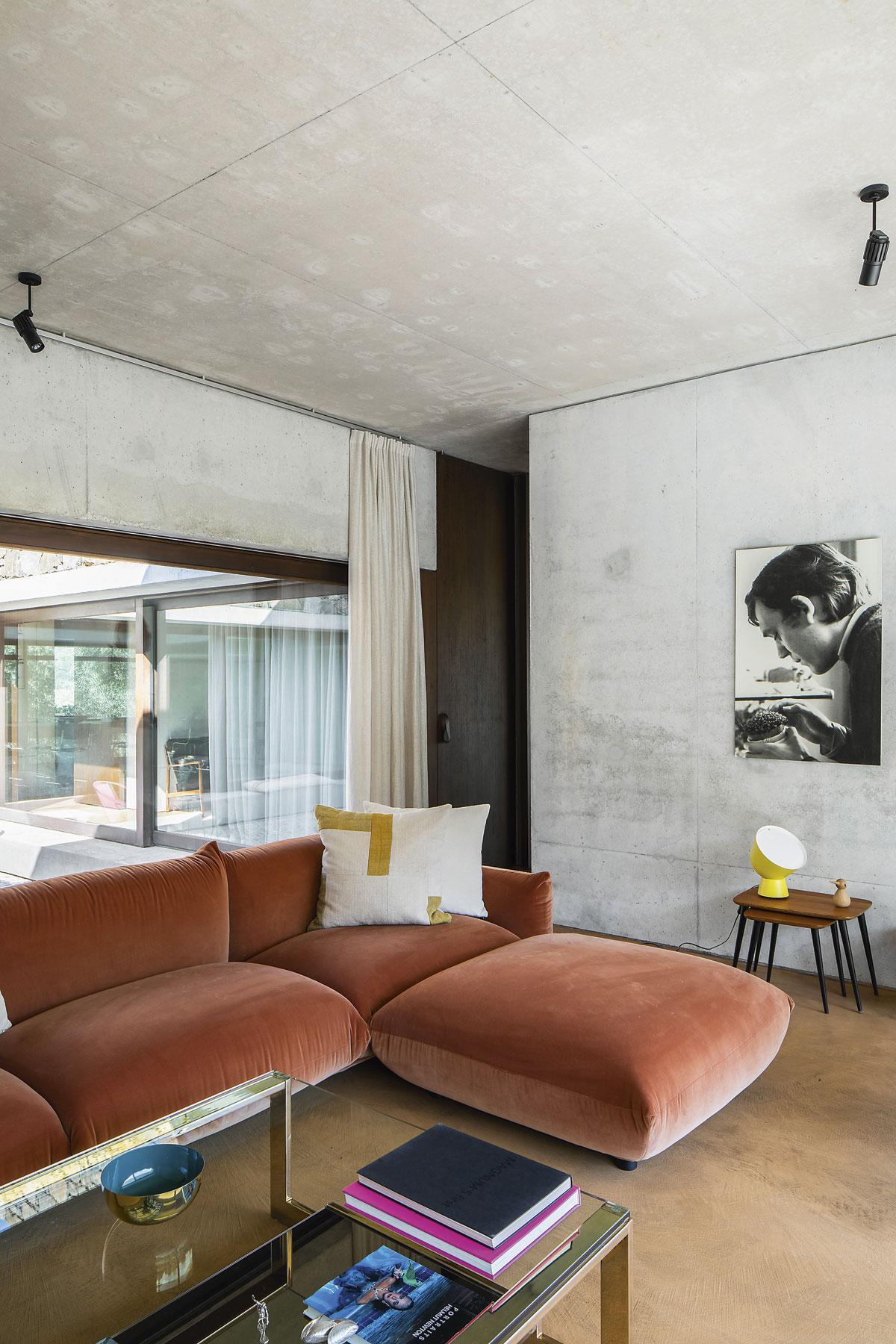 De roze Marenco-sofa (Arflex) en de strogele vloer geven kleur aan het interieur van beton en donker hout. Het gele lampje onder het portret van Jos, de vader van Carlotta, komt uit een PS collectie van IKEA, de salontafel is van Belgo Chrom.