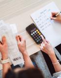 Een foto van twee personen die samen een gezinsbudget opstellen. De ene heeft enkele rekeningen vast, de andere maakt de berekeningen met een rekenmachine op de smartphone.