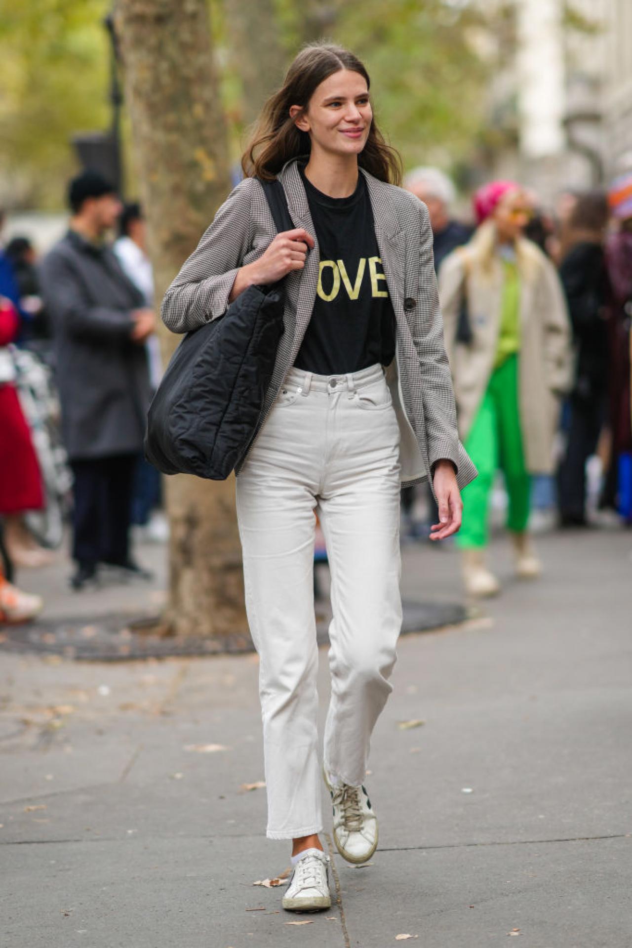 PARIS, FRANCE - OCTOBER 01: A guest wears a black with yellow Love slogan inscription t-shirt, a black and white houndstooth print pattern blazer jacket, a black oversized puffy shoulder bag, white latte high waist denim large pants, white socks, white leather with black V logo sneakers from Veja, outside Loewe, during Paris Fashion Week - Womenswear Spring Summer 2022, on October 01, 2021 in Paris, France. (Photo by Edward Berthelot/Getty Images)