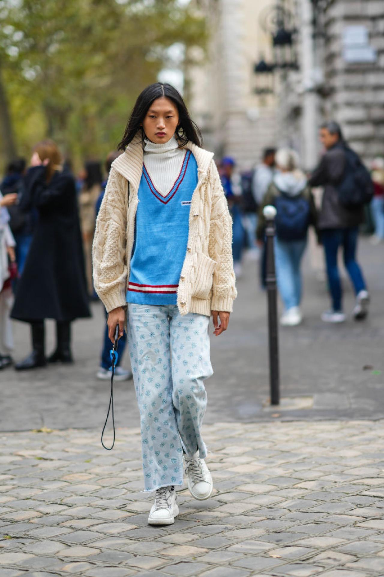 PARIS, FRANCE - OCTOBER 01: A guest wears a white ribbed turtleneck long sleeves t-shirt, a blue wool with red stripes on the collar / V-neck / oversized pullover, a white latte braided wool zipper cardigan, pale blue with blue print flower pattern denim large ripped pants, black and white striped socks, white leather sneakers, outside Loewe, during Paris Fashion Week - Womenswear Spring Summer 2022, on October 01, 2021 in Paris, France. (Photo by Edward Berthelot/Getty Images)