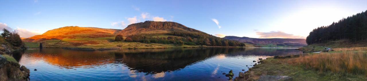 A panorama of Dove Stone Reservoir taken using the new panorama feature and an iPhone 5. Taken just before sunset, the warm light of the sun bathes the surrounding hills in a golden glow. The sky is a deep blue with orange clouds which are both reflected in the water of the reservoir.