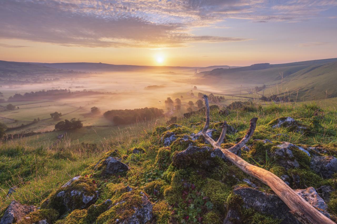 Hope valley on a beautiful misty morning in September 2020 with gritstone rocks and old tree branch in the foreground, Taken at sunrise in the Derbyshire Peak District National park. Castleton, Hope Valley, UK.