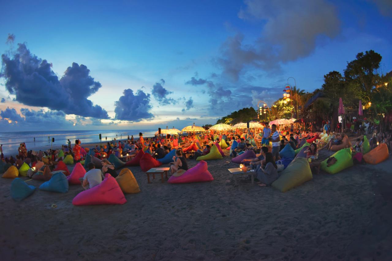 Fisheye view of Double Six beach, with people chilling out at the bars, after sunset. People are having drinks at their puffs, while others walk by the sand. Colourful parasols and lamps lighten the scenery, as the sun casts its last rays of light onto the sky.