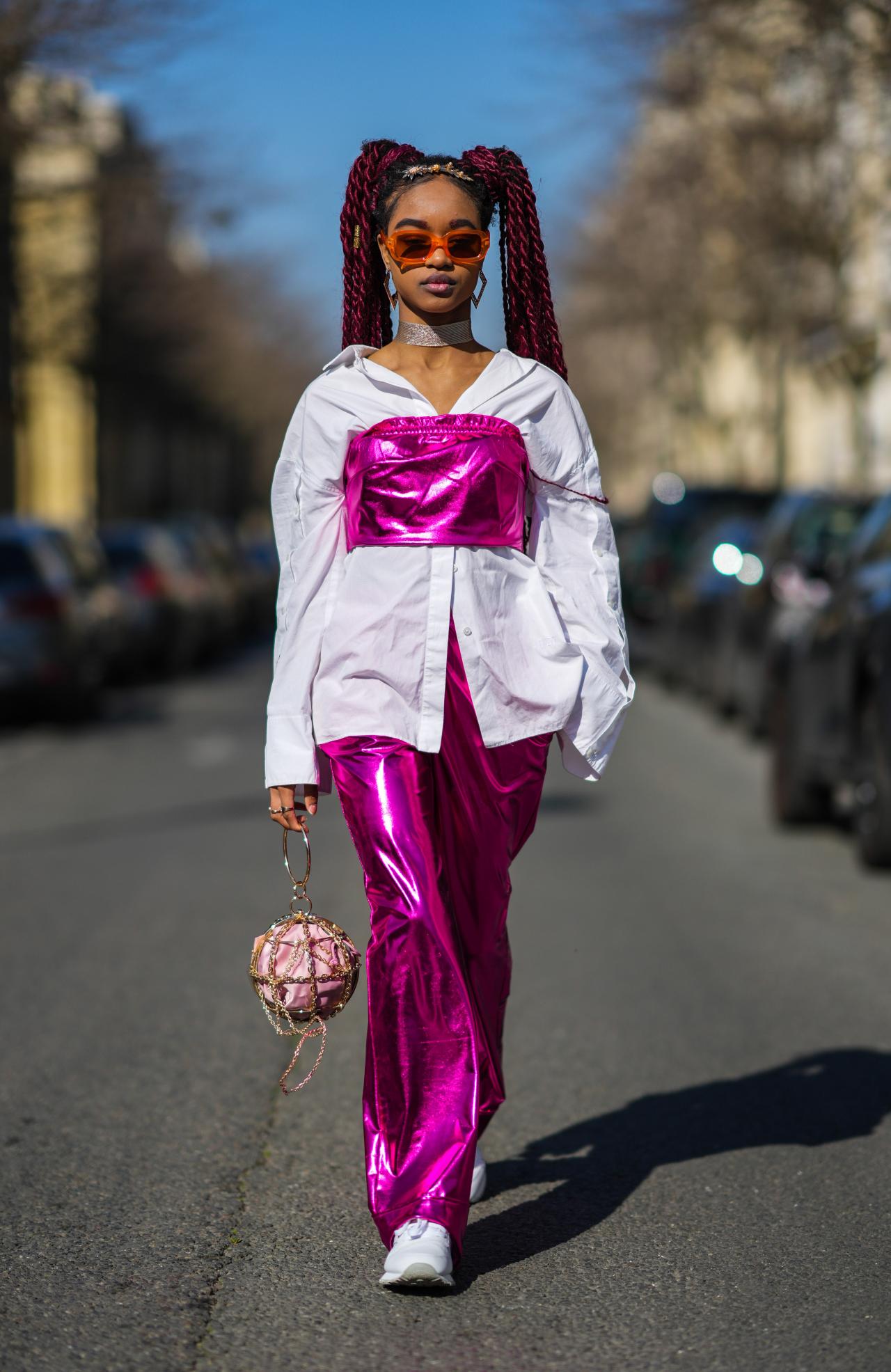 PARIS, FRANCE - MARCH 08: Debbie @blackhey wears a gold flower pattern headband, gold large earrings from Givenchy, orange sunglasses, a white oversized shirt, a neon pink shiny leather shoulder-off / cropped top, matching neon pink shiny leather large pants, a gold chain circle handbag with a pale purple clutch inside, white leather sneakers, outside Chanel, during Paris Fashion Week - Womenswear F/W 2022-2023, on March 08, 2022 in Paris, France. (Photo by Edward Berthelot/Getty Images)