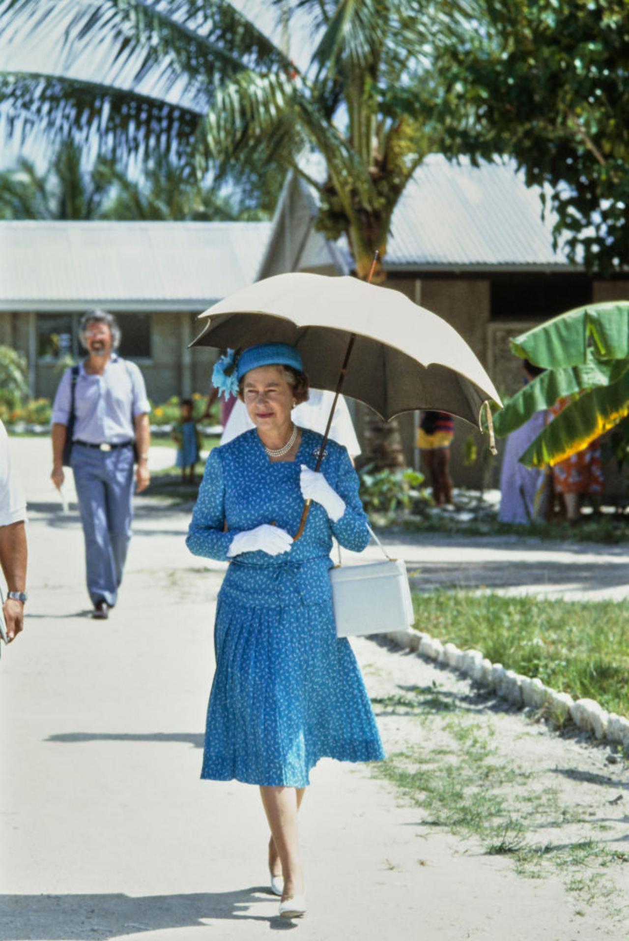 British Royal Queen Elizabeth II, wearing a blue two-piece outfit with white detail and a matching hat, shades herself beneath a parasol during a visit to the Princess Margaret Hospital in Fongafale, on the South Pacific of Tuvalu, 27th October 1982. The visit was part of the Royal Tour of Australia and the Pacific Islands. (Photo by Tim Graham Photo Library via Getty Images)