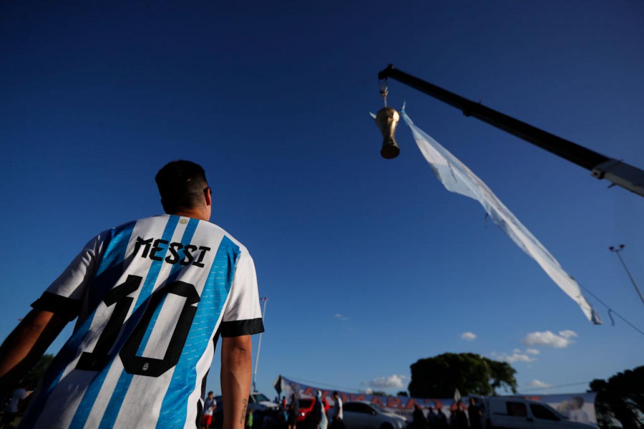 BUENOS AIRES, ARGENTINA - DECEMBER 19: A fan wears a jersey of Lionel Messi watches a large replica of the FIFA World Cup hanging from a crane before the arrival of the Argentina men's national football team after winning the FIFA World Cup Qatar 2022 on December 19, 2022 in Buenos Aires, Argentina. (Photo by Marcos Brindicci/Getty Images)
