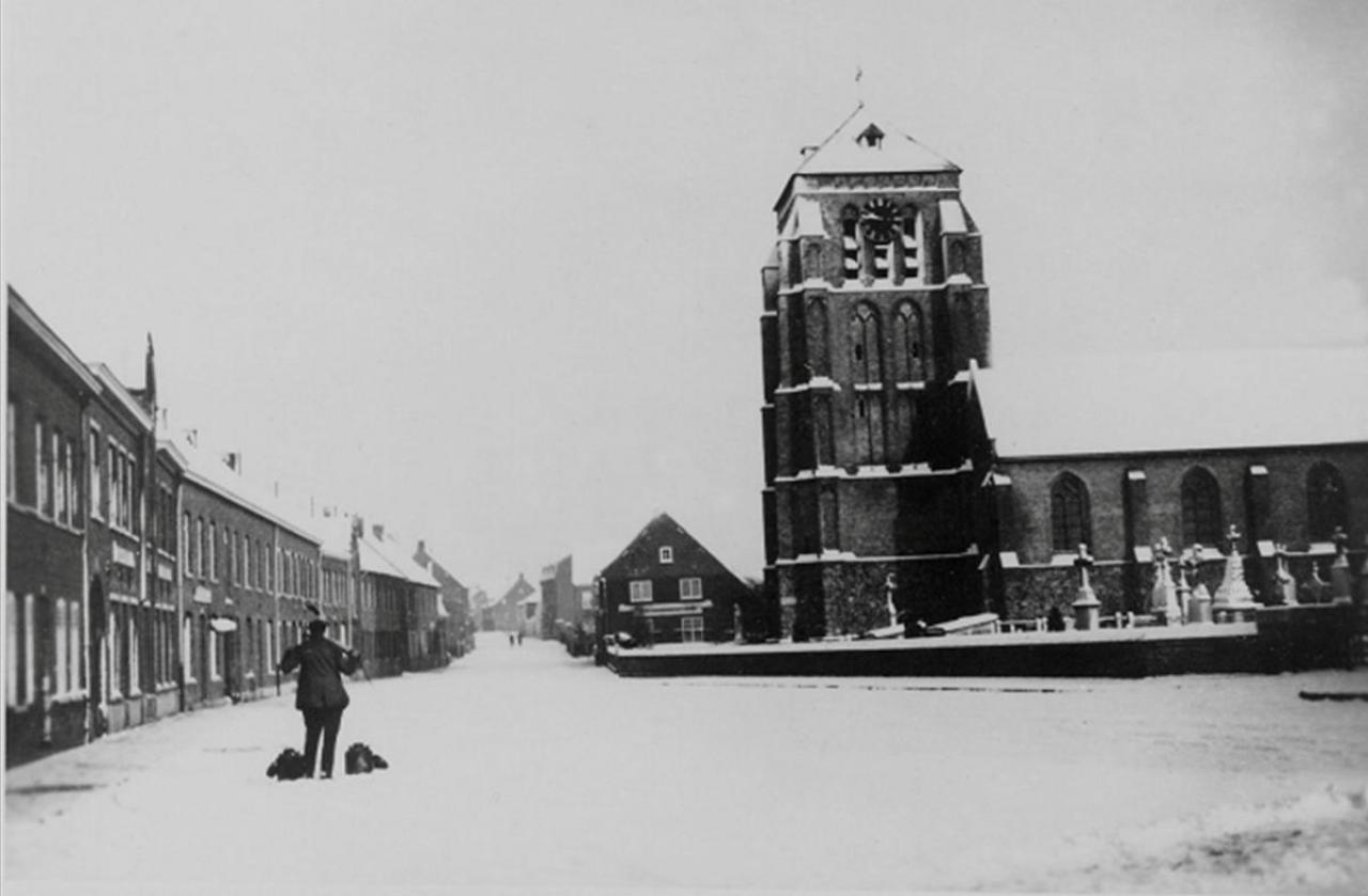 Zo te zien was er in de winter van 1930 nogal wat sneeuw gevallen. Op de verlaten dorpsplaats loop Jozef Deberdt met zijn juk en petroleum ketels om zijn klanten van brandstof voor hun lampen en petroleumvuurtjes te voorzien.