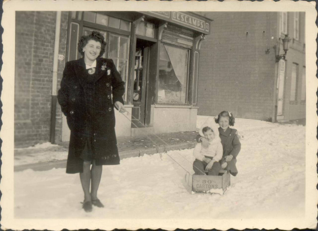 Sneeuwpret in de Pennestraat rond 1940. Leonie Clarysse trekt haar kinderen Roland en Huguette door de sneeuw met een zelfgemaakte slede. Achterzin zie je de winkel die zij samen met haar man Fernand Coffyn uitbaatte.