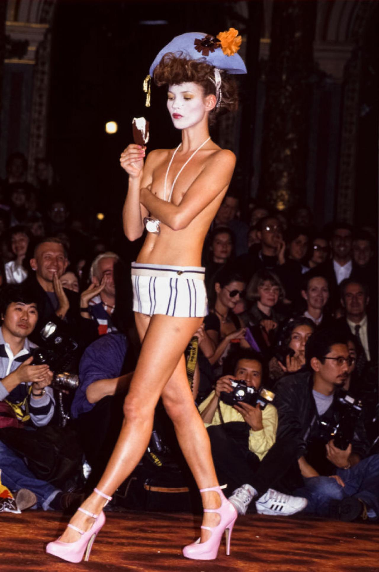 Kate Moss tijdens de show van Vivienne Westwood in 1993. Topless eet het model een ijsje terwijl ze over de catwalk loopt.