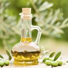 Flesje olijfolie omringd door onbewerkte olijven buiten op een tafel met olijfboom op achtergrond.