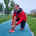 Oudere man van in de zestig knielt op een blauwe atletiekbaan in een park en houdt zijn enkel vast van de pijn na het joggen. Dit beeldt een sportblessure, overbelasting en een ongeluk tijdens het sporten in de buitenlucht uit.