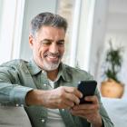 Grijze man van middelbare leeftijd zit in de zetel terwijl hij zijn smartphone gebruikt.