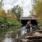 Artiest met fototoestel langs de kant van een rivier in stadsgedeelte met groen.