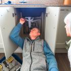 Een loodgieter inspecteert de leidingen onder de gootsteen in de keuken, terwijl de huiseigenaar toekijkt.