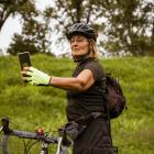 Sportieve oudere vrouw op de fiets die buiten op een pad in een groen park haar smartphone gebruikt.