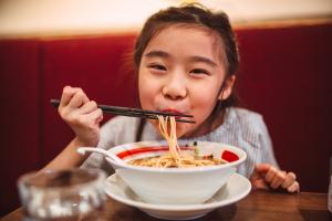 Lovely cheerful girl smiling joyfully at camera while enjoying noodle soup in restaurant.