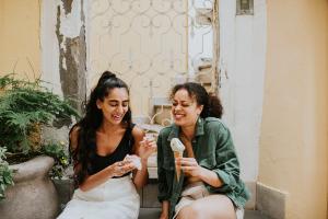 Two friends sit on a step in a European alleyway. They enjoy an ice cream together. One eats from a tub, and the other holds a cone. They giggle as one of the woman has some strawberry ice cream on her nose.