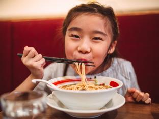 Lovely cheerful girl smiling joyfully at camera while enjoying noodle soup in restaurant.