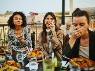 Medium shot of women laughing and savoring traditional Moroccan tajine at scenic rooftop eatery during holiday in Marrakech