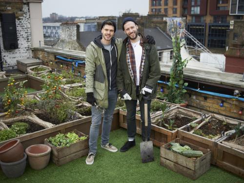Two young men smiling straight to camera in urban allotment on roof garden, holding a spade and a gardening fork.