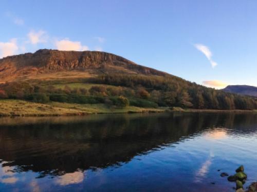 A panorama of Dove Stone Reservoir taken using the new panorama feature and an iPhone 5. Taken just before sunset, the warm light of the sun bathes the surrounding hills in a golden glow. The sky is a deep blue with orange clouds which are both reflected in the water of the reservoir.