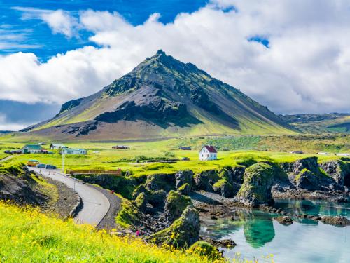 View of the basalt rocks formation on the coastline with the beautiful houses and the Mountain Stapafell in the background at Arnarstapi Village in summer sunny day in Iceland