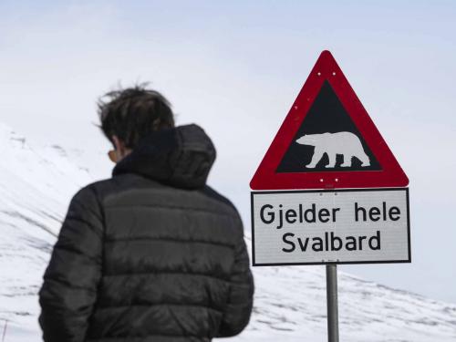 A man is pictured standing next to a warning sign depicting a polar bear at the side of a road near Longyearbyen on May 2, 2022, on the Svalbard Archipelago, northern Norway. - Home to polar bears, the midnight sun and the northern lights, a Norwegian archipelago perched high in the Arctic is trying to find a way to profit from its pristine wilderness without ruining it. The Svalbard archipelago, located 1,300 kilometres (800 miles) from the North Pole and reachable by commercial airline flights, offers visitors vast expanses of untouched nature, with majestic mountains, glaciers and frozen fjords. Or, the fjords used to be frozen. Svalbard is now on the frontline of climate change, with the Arctic warming three times faster than the planet. (Photo by Jonathan NACKSTRAND / AFP)