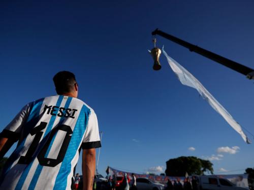 BUENOS AIRES, ARGENTINA - DECEMBER 19: A fan wears a jersey of Lionel Messi watches a large replica of the FIFA World Cup hanging from a crane before the arrival of the Argentina men's national football team after winning the FIFA World Cup Qatar 2022 on December 19, 2022 in Buenos Aires, Argentina. (Photo by Marcos Brindicci/Getty Images)