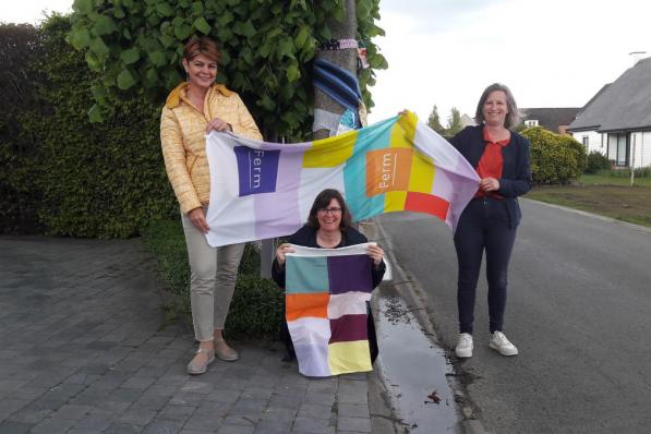 Greetje Lisabeth, Nancy Vanelverdinghe en Els Decock tonen de kleurrijke stroken brei-, haak- en naaiwerk dat in de buurt van de locaties hangt.© TV