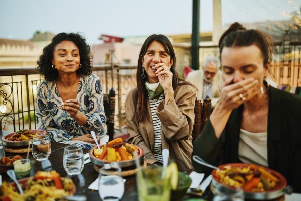 Medium shot of women laughing and savoring traditional Moroccan tajine at scenic rooftop eatery during holiday in Marrakech