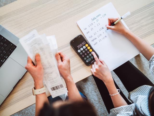Een foto van twee personen die samen een gezinsbudget opstellen. De ene heeft enkele rekeningen vast, de andere maakt de berekeningen met een rekenmachine op de smartphone.