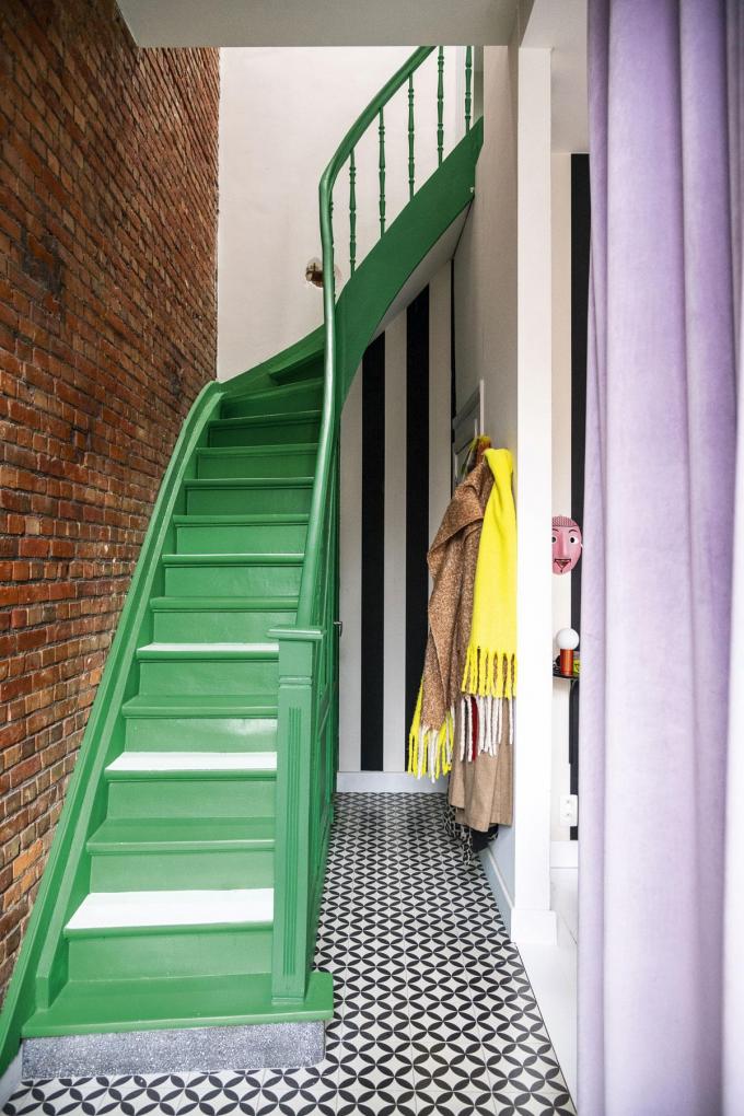 de trap is groen en wit geschilderd. Het zware gordijn tussen de traphal en de woonkamer vervangt een tussenmuur en is lila aan de ene kant – past mooi bij het groen van de trap, vindt Jasmien – en spiegelgoud aan de andere kant. Jasmien zocht heel lang naar de juiste stoffen.