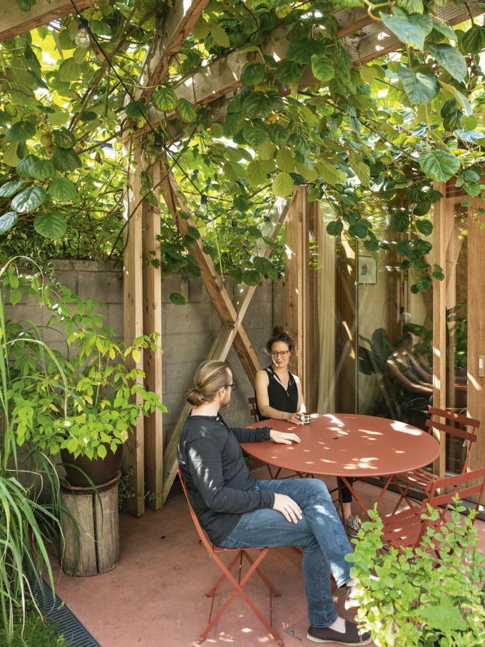 De structuur van constructiehout start in de keuken en loopt de hele woonkamer door tot in de tuin, waar de kiwiplant van de buren schaduw brengt. Het houten patroon verlengt de ruimte visueel, de Dibond-platen op de tuinberging zorgen voor een optische illusie.