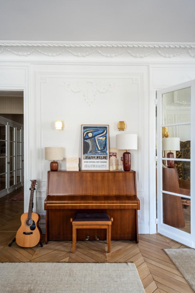 De vintage teak piano en gitaar vormen een muzikale hoek in de woonkamer. De twee Traviata-lampen zijn van Les Composantes, de nieuwe interieurcollectie van Sézane.