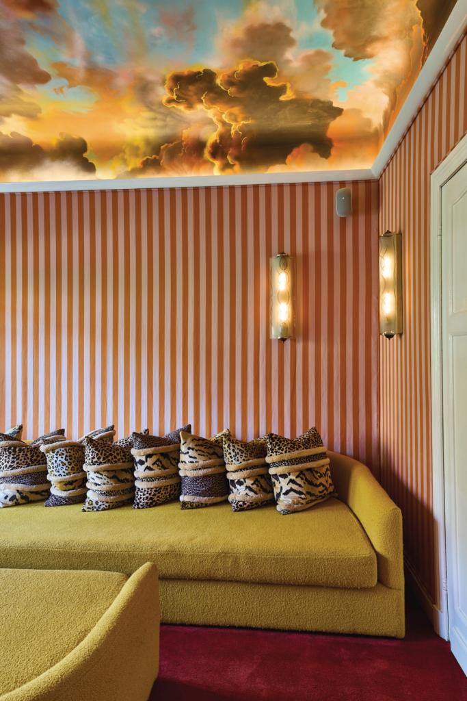 Tv-kamer in Brugge. ‘Het plafond is geschilderd door een graffitikunstenaar wiens werk ik had gespot onder een brug. Na een lange zoektocht kon ik hem opsporen dankzij zijn initialen. Het is een moderne versie van de achttiende-eeuwse Italiaanse en Franse wolkenplafonds, maar dan met een paar donkere wolken om het toch wat te doen schuren. De gang met zicht op de muziekkamer komt uit dezelfde woning. Het ontbrak haar oorspronkelijk aan eenheid. Met de juiste kleuren maakten we er een geheel van.’